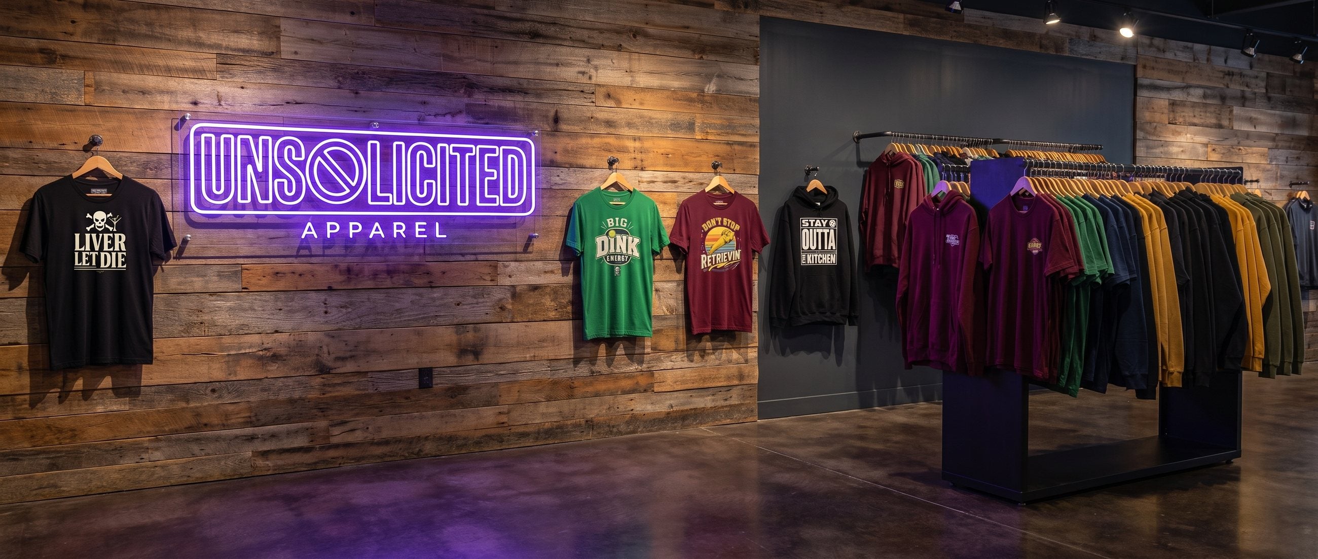 The Unsolicited Apparel shop wall: a violet neon Unsolicited Apparel sign on reclaimed wood above graphic tees and a hoodie, with a full rack of shirts alongside.